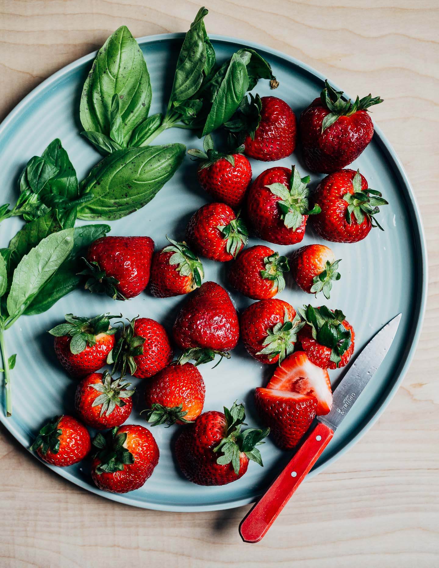 A light blue platter with strawberries, fresh basil leaves, and a red paring knife. 