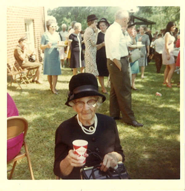 A nicely dressed lady sitting down at a church picnic. People are milling around in the background. 