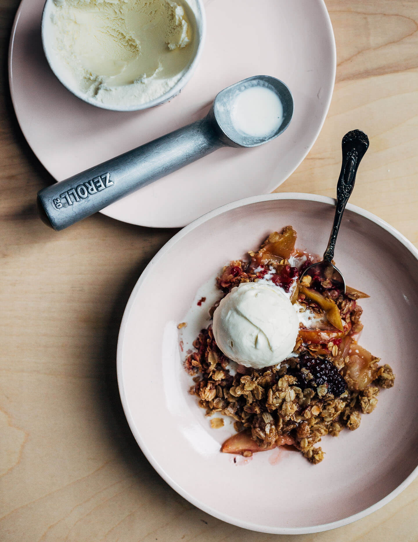 A pink bowl with a serving of crisp and a scoop of ice cream. Another plate holds a tub of ice cream and an ice cream scoop. 