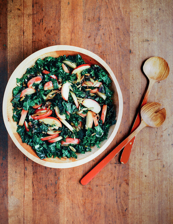 A bowl of kale salad on a wooden table with salad tongs alongside.