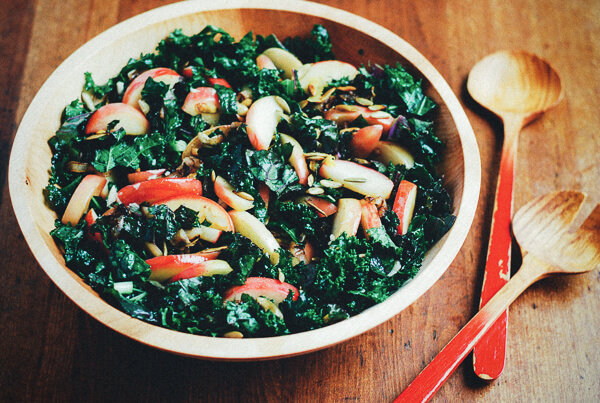 A bowl of kale salad on a wooden table with salad tongs alongside. 