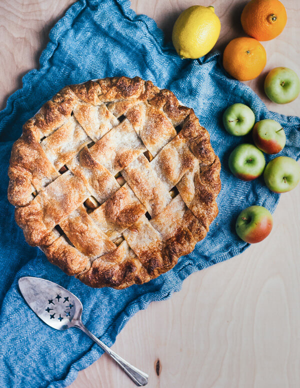 A lattice-top pie on a dish cloth. There’s a serving utensil, citrus fruits, and lady apples alongside. 