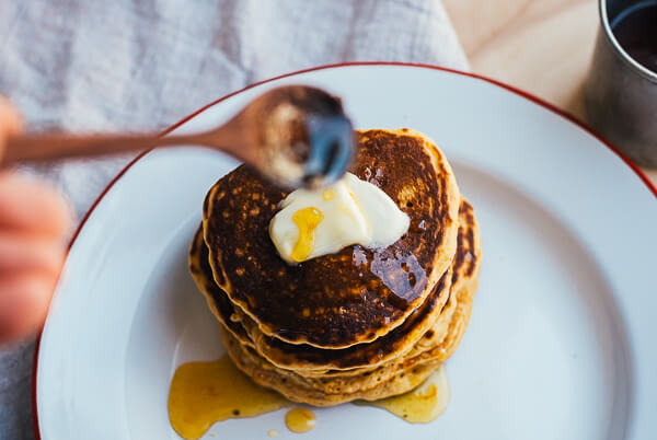 A hand is spooning syrup over a stack of pancakes with butter. 