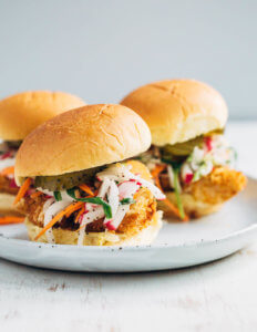 A plate with three fish sandwiches layered with pickles, and radish and turnip slaw
