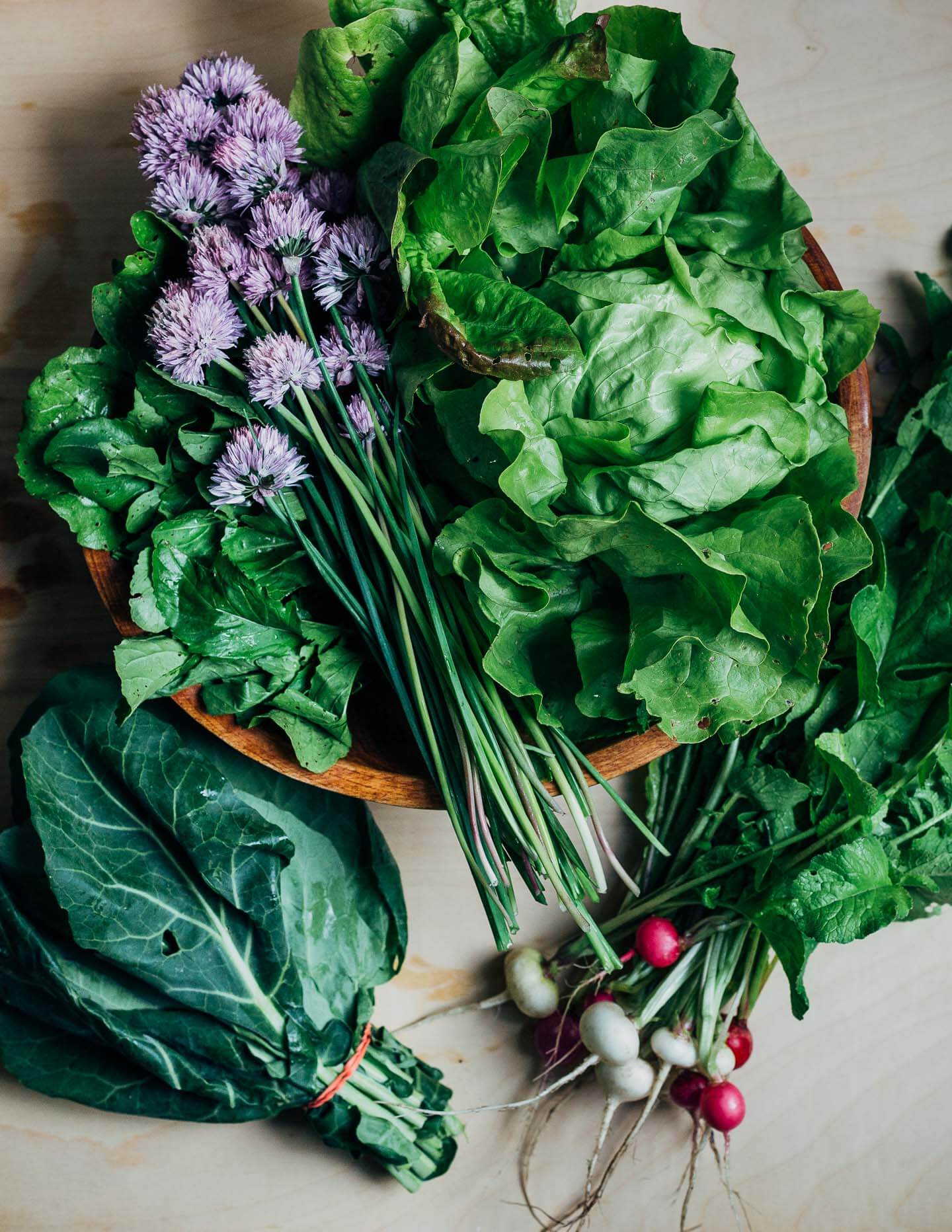 Spring vegetables laid out and in a bowl, with radishes, lettuce, collard greens and chive blossoms. 