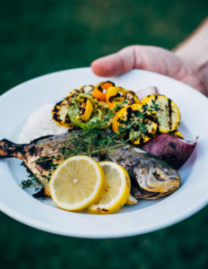 A hand holding out a plate of grilled fish and vegetables