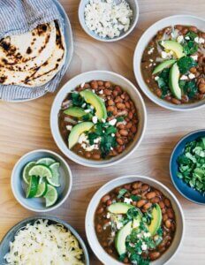 Bowls of bean, tortillas, sliced avocados, cilantro, and other fixings