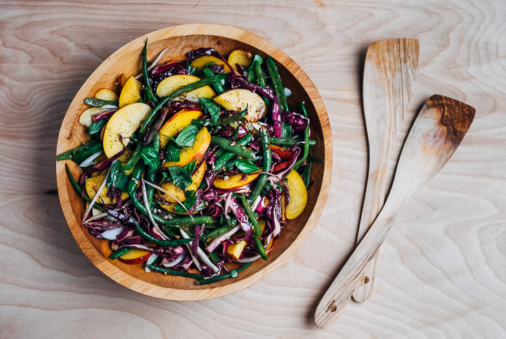 A green bean and peach salad in a wooden bowl with wooden salad tongs alongside. 