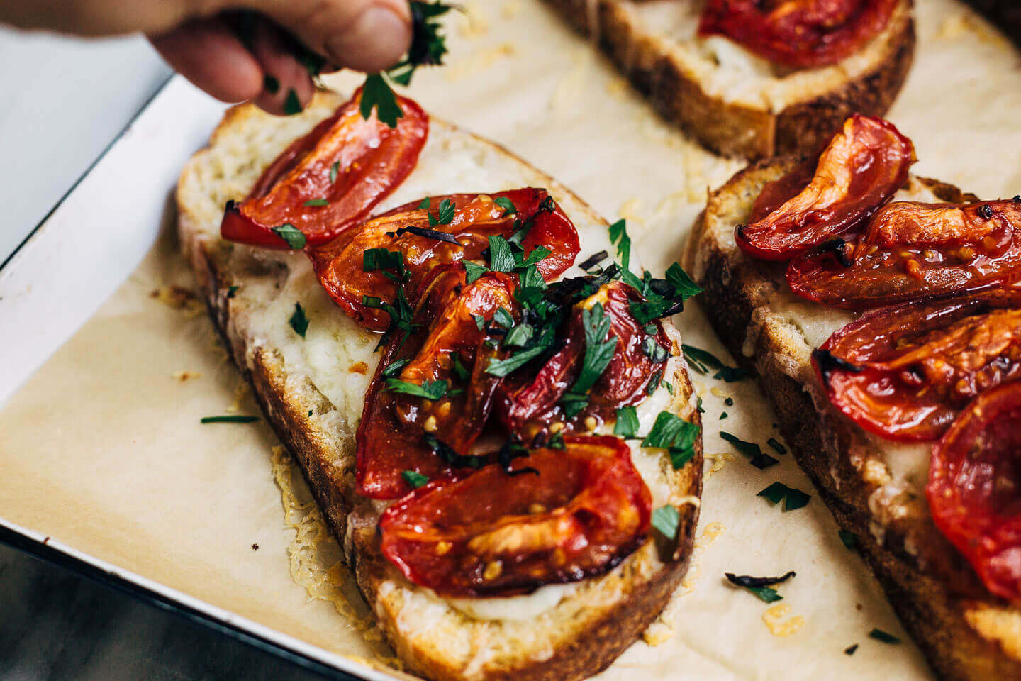 A tartine with melted cheese and roasted Roma tomatoes. A hand is placing chopped basil on top. 