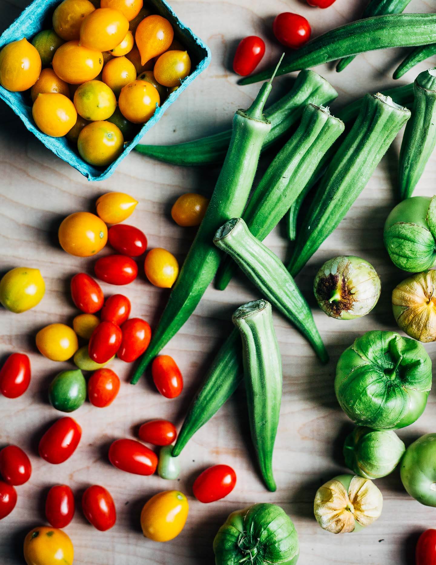 An assortment of red and yellow cherry tomatoes, okra, and tomatillos on a table. 