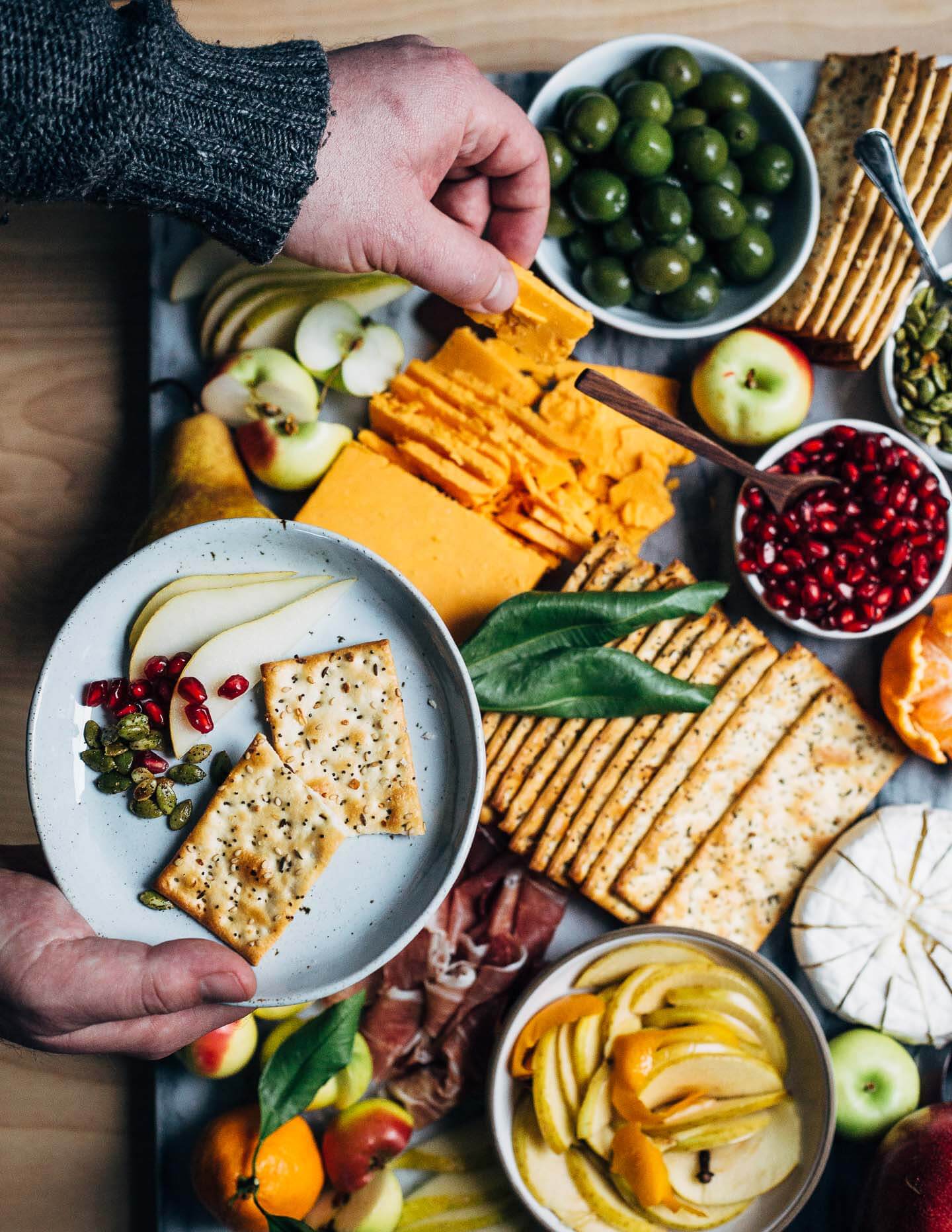 A modern holiday cheese board with red cheddar and brie-style cheeses, winter fruit, and sweet and sour quick-pickled apples.