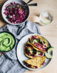 Spicy salmon tacos make for a delicious springtime feast. Tender, crisp skinned salmon is suffused with chipotle chili powder, cumin, and coriander, tucked into toothsome corn tortillas, and layered with a fennel and red cabbage slaw and an easy cilantro-avocado blender sauce. 