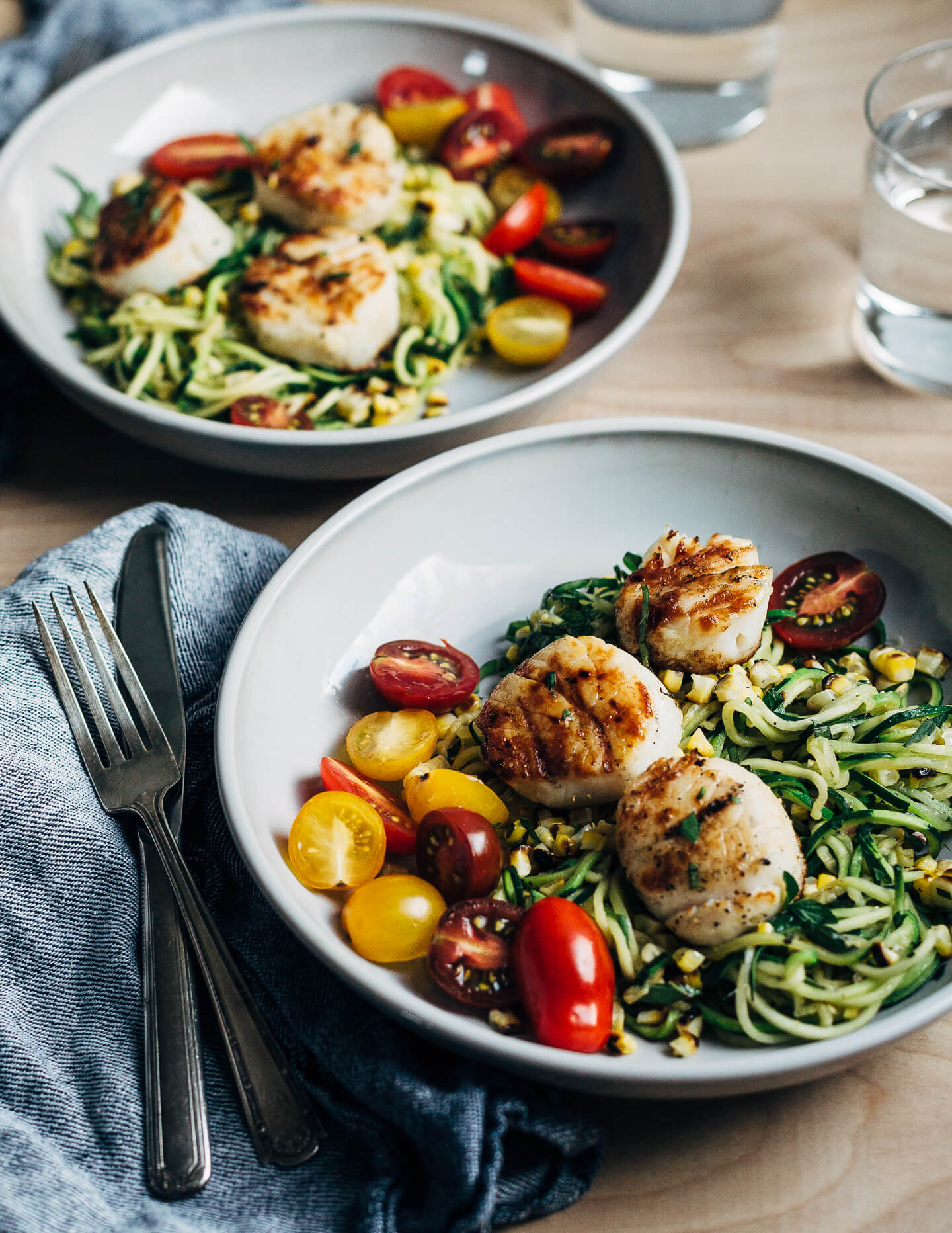 A simple grilled summer feast featuring grilled scallops served atop zucchini noodles tossed with charred corn and fresh herbs.