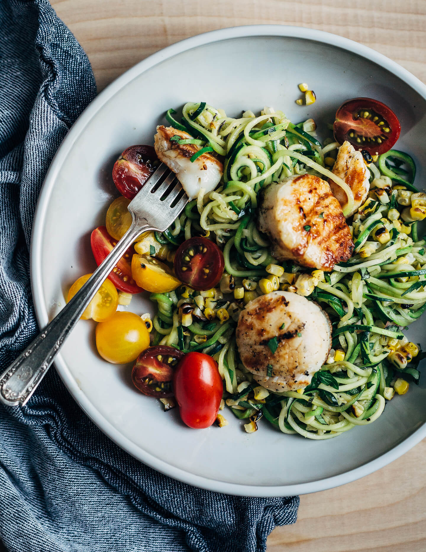 A simple grilled summer feast featuring grilled scallops served atop zucchini noodles tossed with charred corn and fresh herbs.