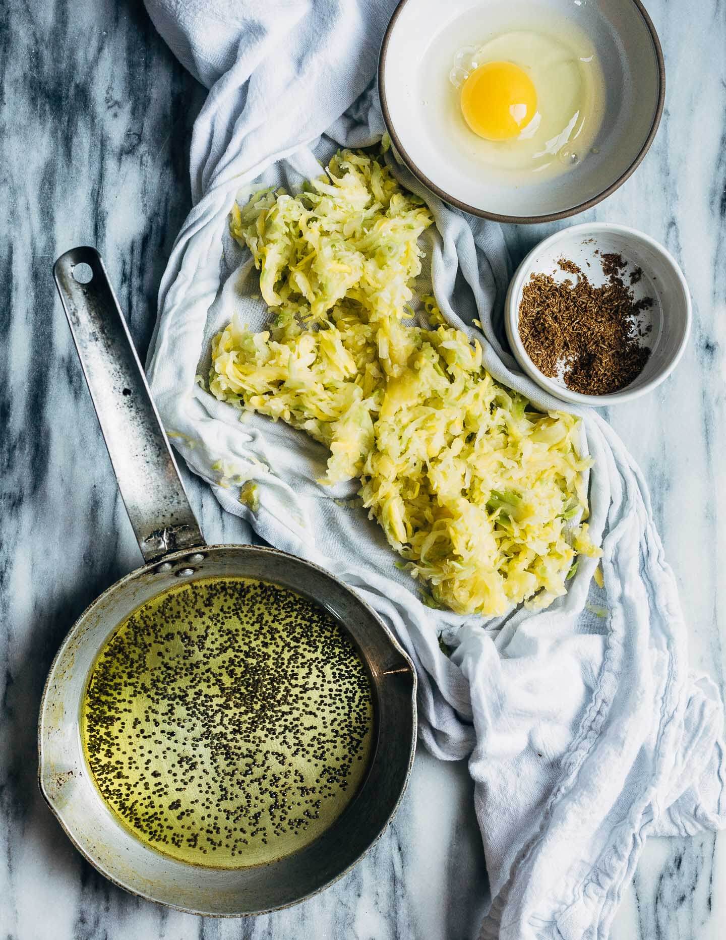A work surface with shredded squash in a cheese cloth and fried mustard seeds in a small pan, plus a bowl with an egg and one with ground spices. 