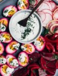 Beet pickled eggs on a platter with a creamy sauce, beet pickles, and herbs.