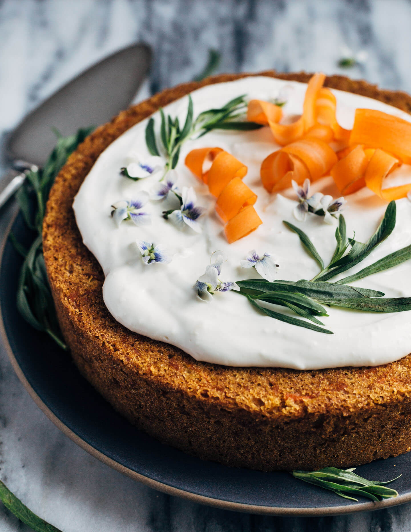 A simple olive oil carrot cake for spring celebrations! The tender, springy whole wheat crumb is suffused with nutty olive oil, fresh carrots, and orange zest. The cake is topped with an airy, lightly sweet yogurt and cream cheese frosting, carrot ribbons, tarragon, and edible flowers. 