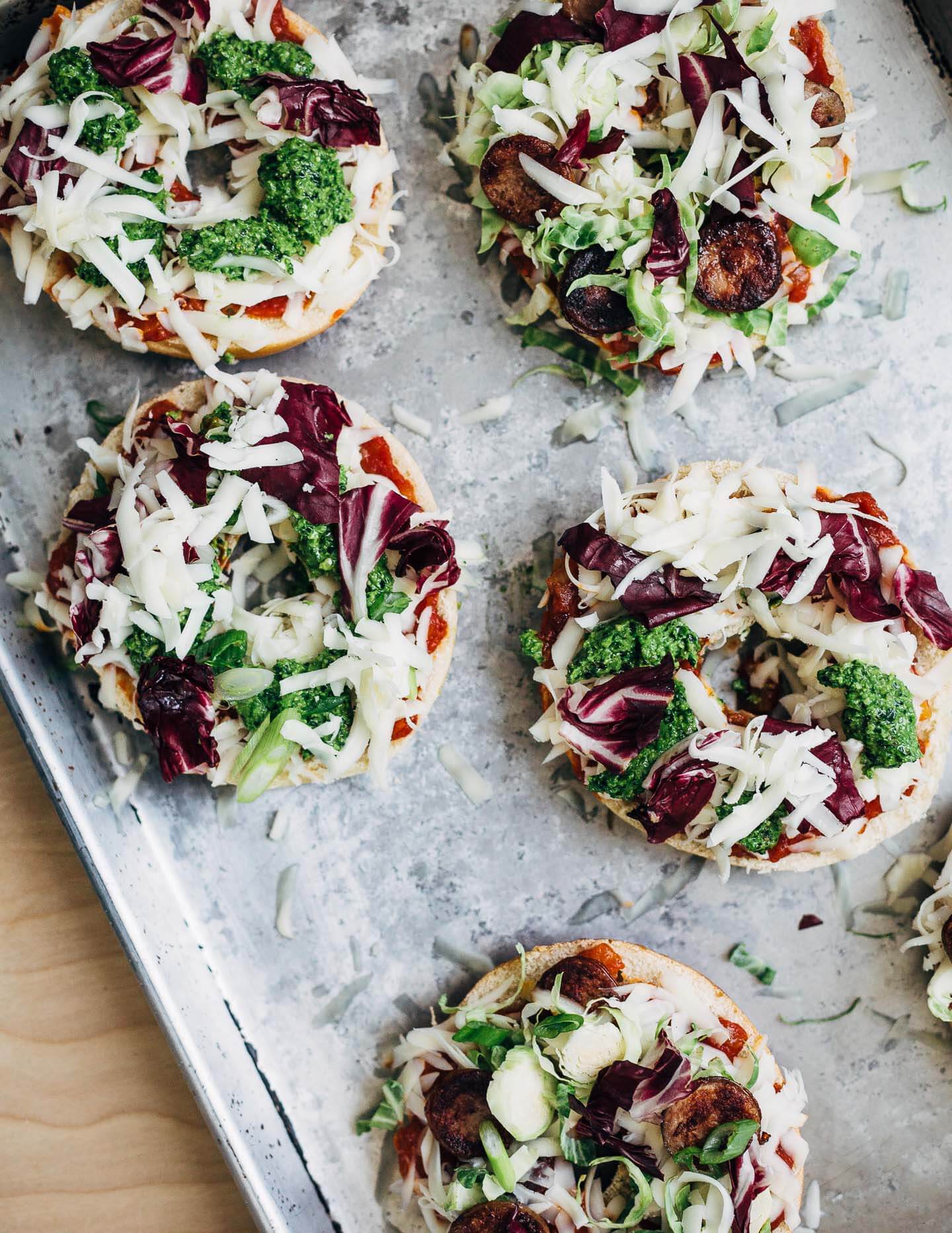 Pizza bagels with pesto, sausage, and spring vegetables, ready for the oven.