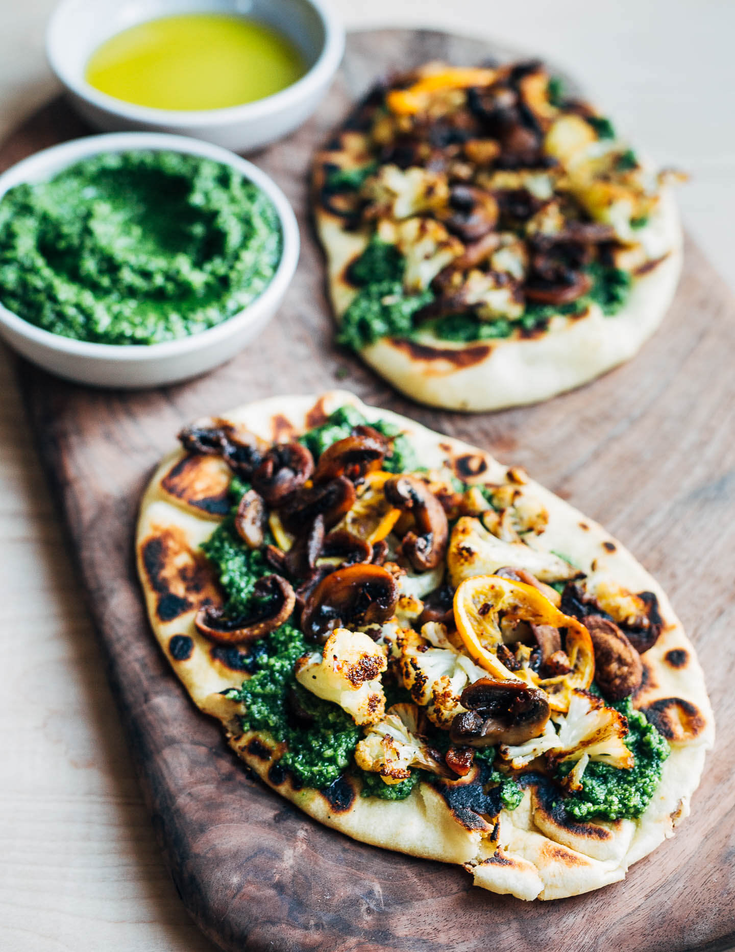 Finished roasted cauliflower flatbreads. The flatbreads are arranged on a cutting board along with little bowls of pesto and olive oil. 
