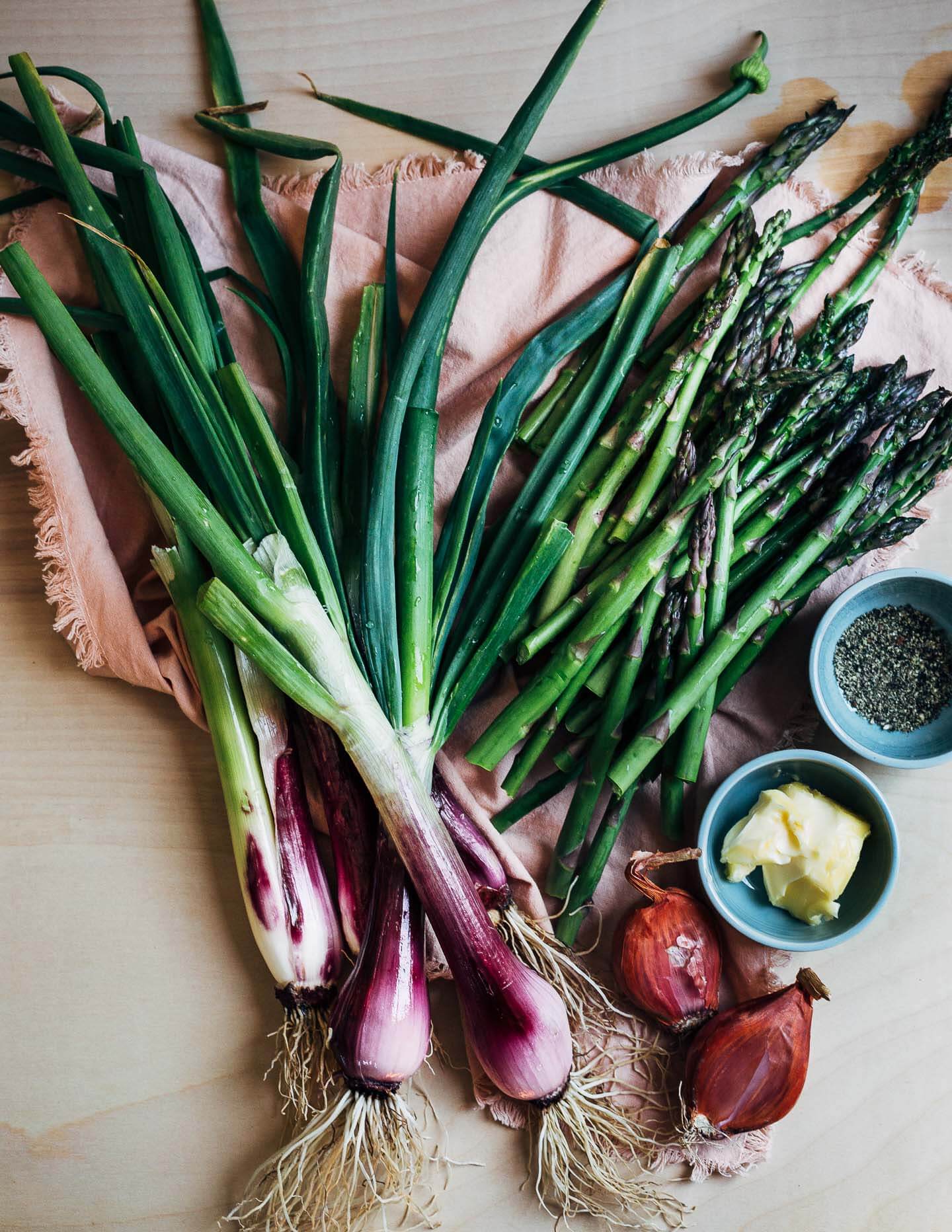 A flatlay with spring onions, asparagus, and shallots, plus little bowls with butter and black pepper