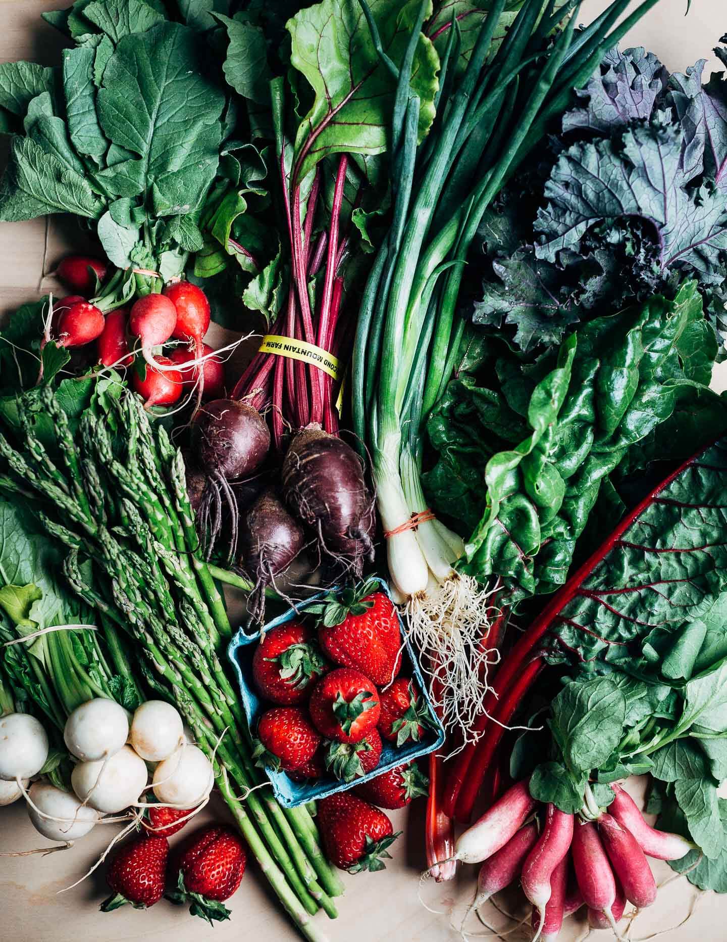 A table piled with spring vegetables, including asparagus, beets, greens, and strawberries. 