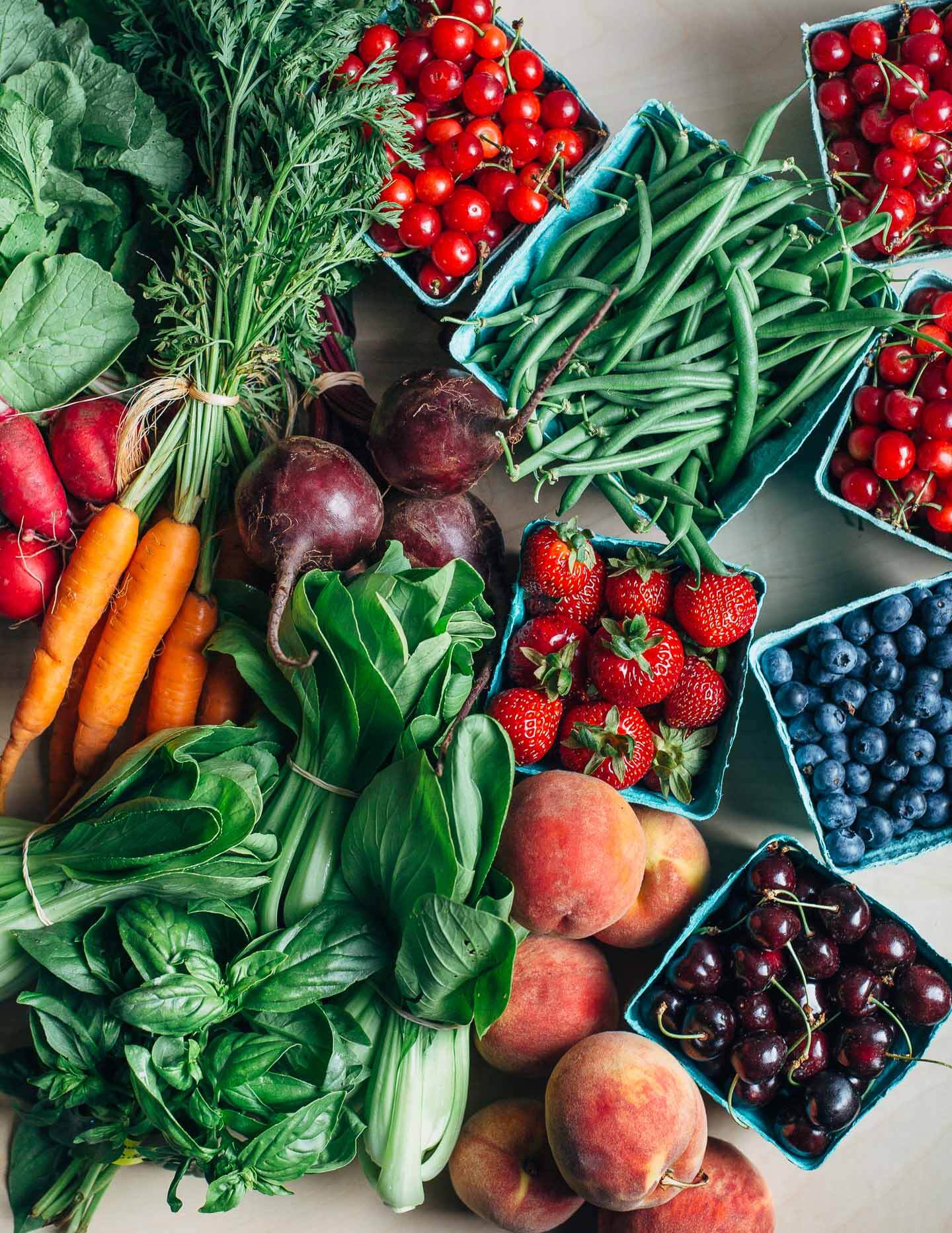 Produce, including radishes, carrots, beets, basil, bok choy, peaches, strawberries, cherries, and blueberries, arranged on a tabletop.