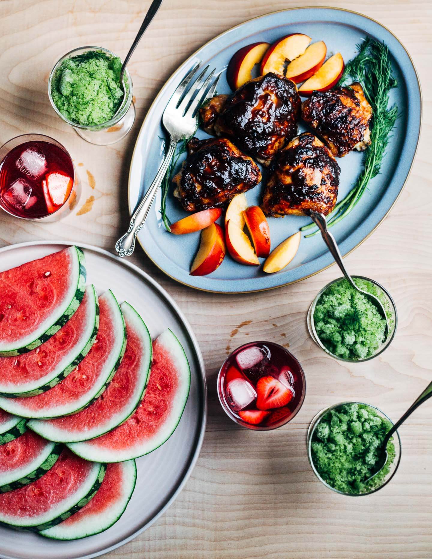 A spread of dishes and platters of food, with chicken, watermelon, drinks, and granita.