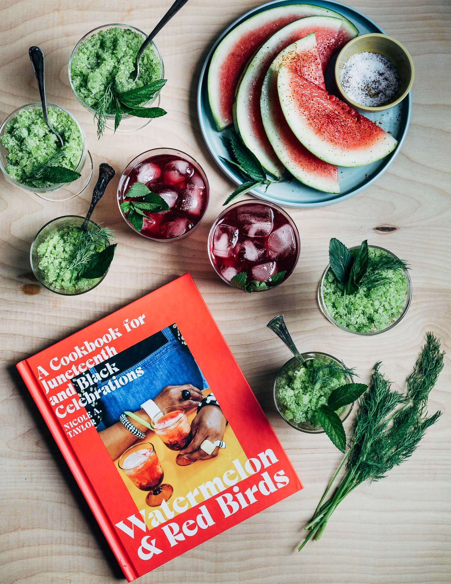 The cookbook Watermelons and Red Birds alongside glass of hibiscus tea, cucumber granita, and sliced watermelon.