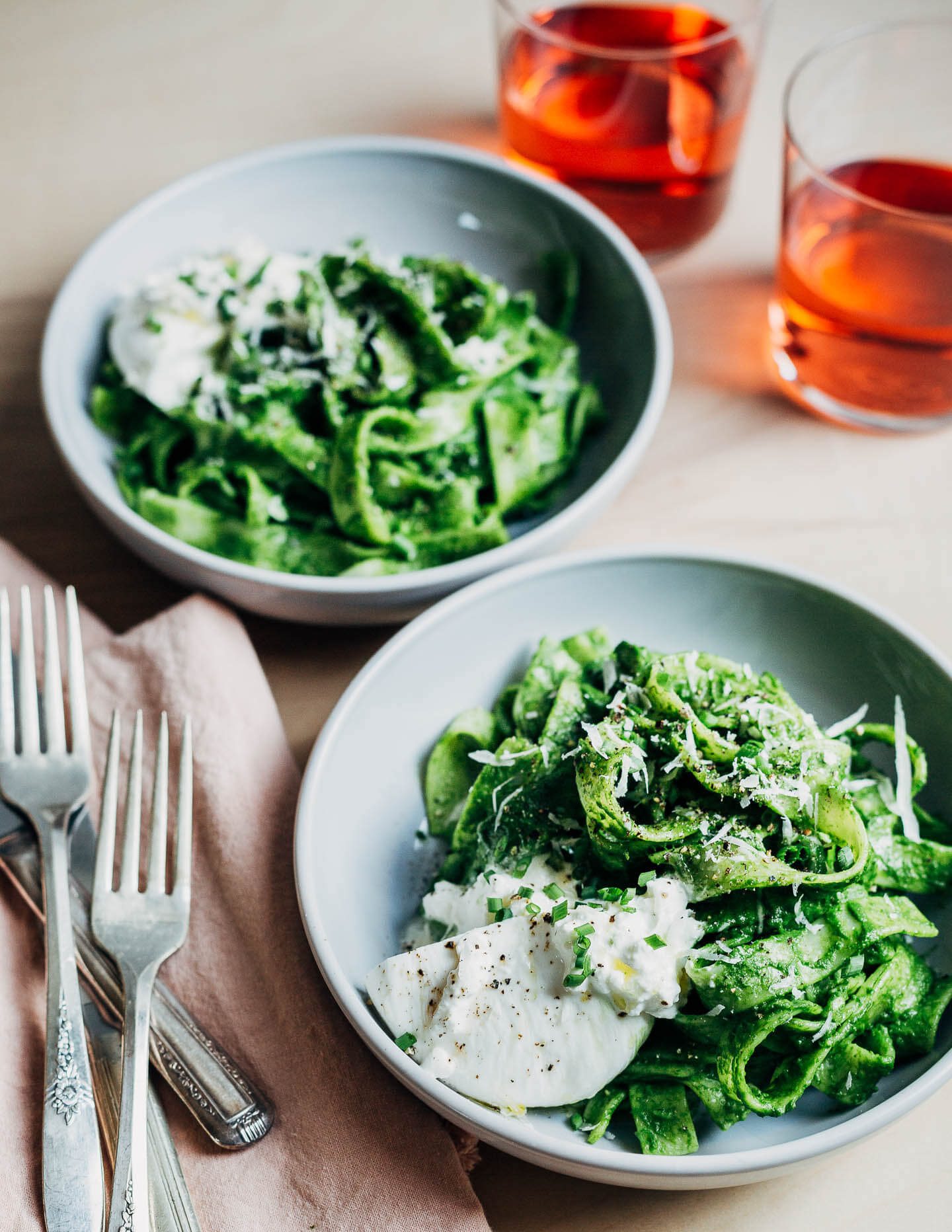 Two bowls of bright green pasta noodles with drinks and utensils on the side.