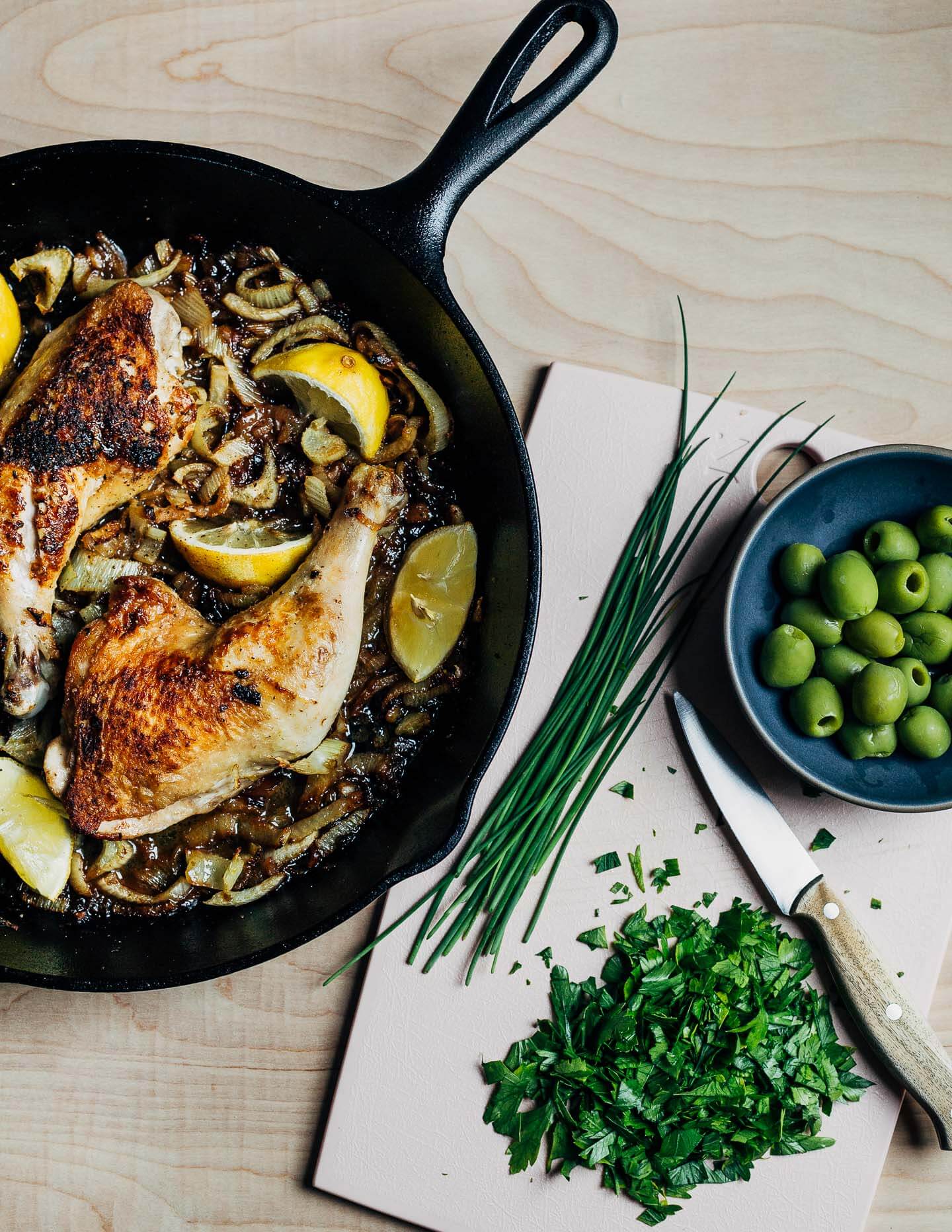 A cast iron skillet with roasted chicken and a cutting board with herbs.