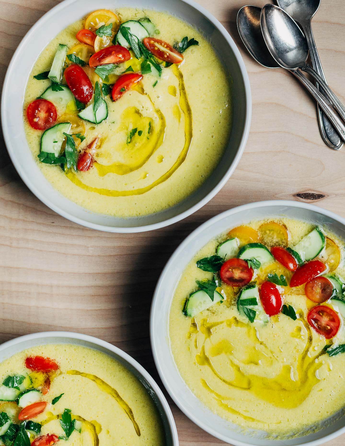 Three bowls of yellow gazpacho garnished with cherry tomatoes, cucumbers, and herbs.