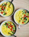 Three bowls of yellow gazpacho garnished with cherry tomatoes, cucumbers, and herbs.