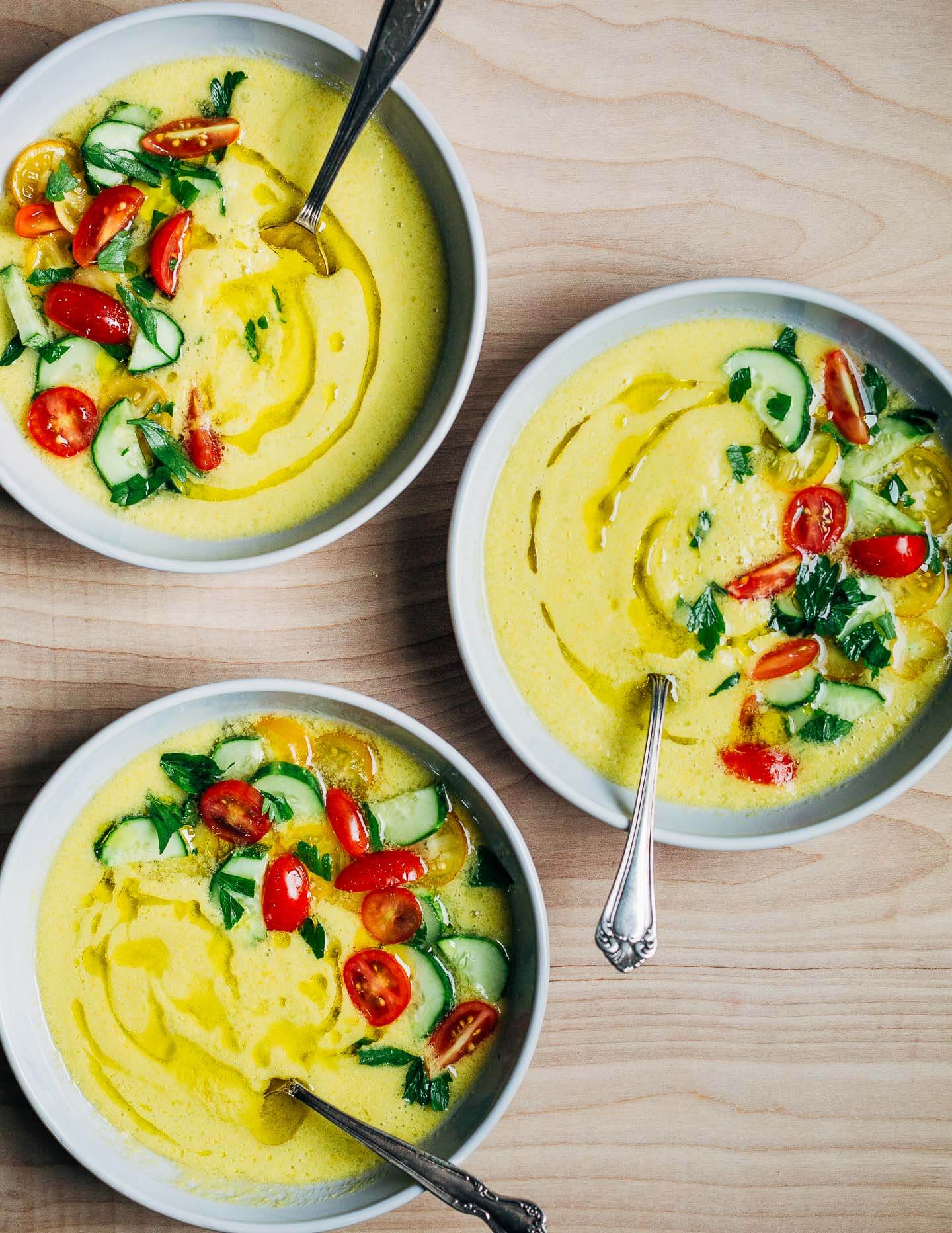 Three bowls of yellow gazpacho garnished with cherry tomatoes, cucumbers, and herbs. 