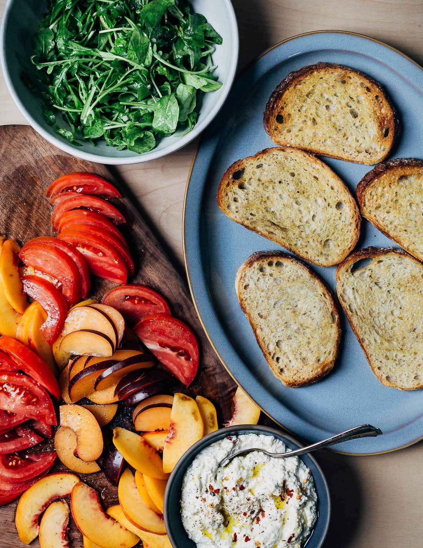 All the ingredients to assemble ricotta toasts, including crostini, arugula, sliced fruit, and ricotta. 