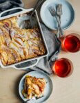 A tabletop with a custard cake with a slice spooned out, glasses of cider, and a copy of the 'Everyday Cake' cookbook.