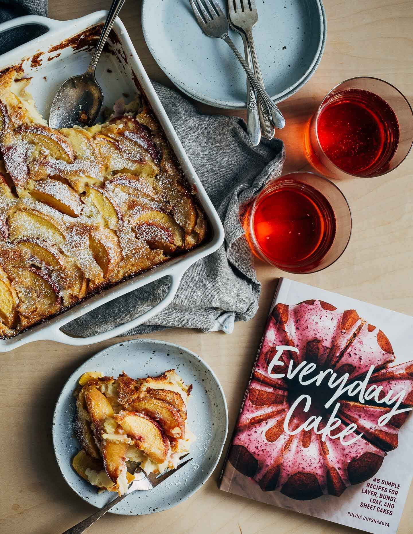 A scene with a custard cake, a slice of cake, plates, forks, cider in glasses, and a copy of the 'Everyday Cake' cookbook. 