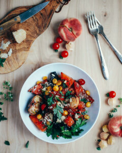 A colorful watermelon and peach salad on a table, with utensils, cheese, and tomatoes scattered all around.