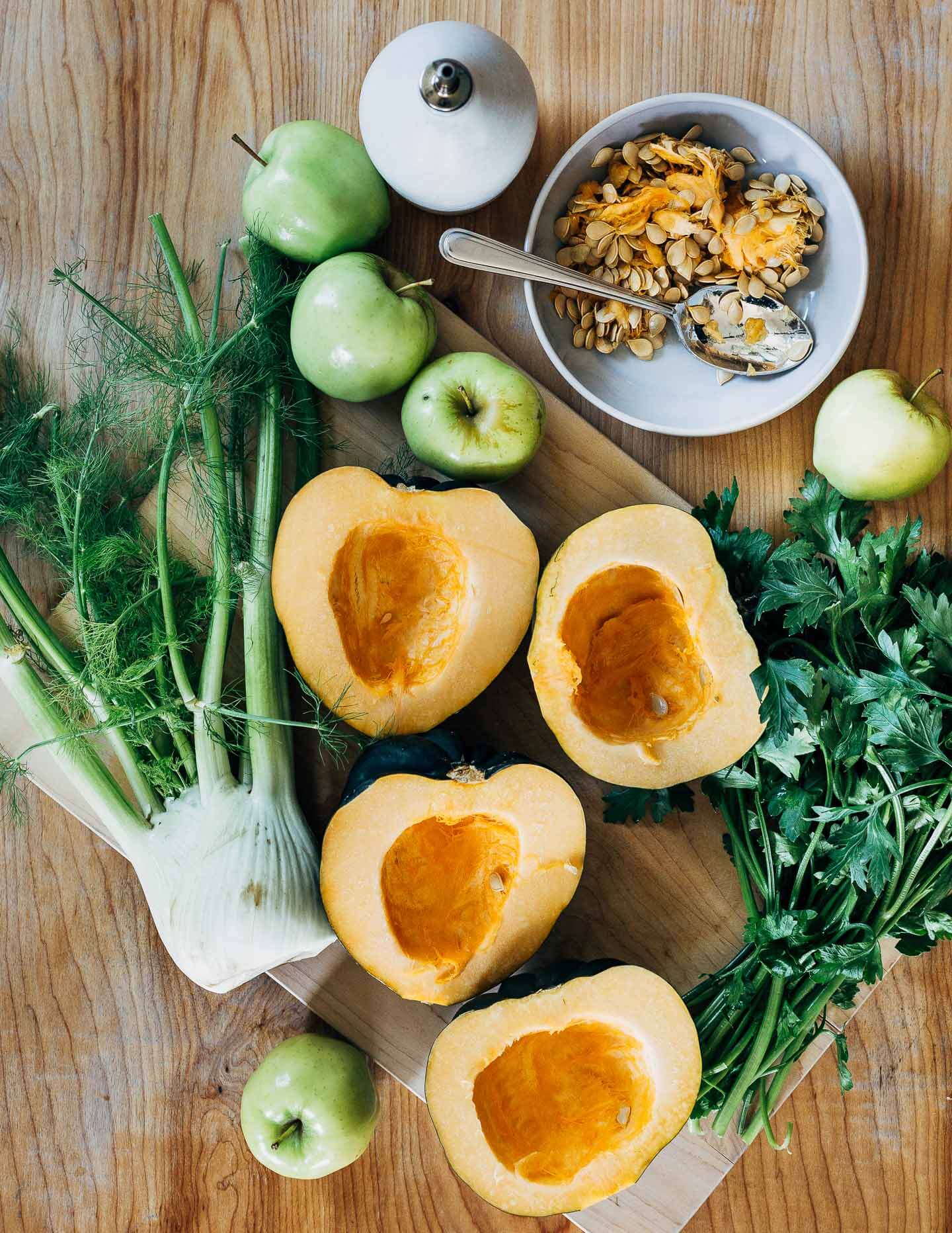 A cutting board with fennel, acorn squash halves, parsley, and apples. 