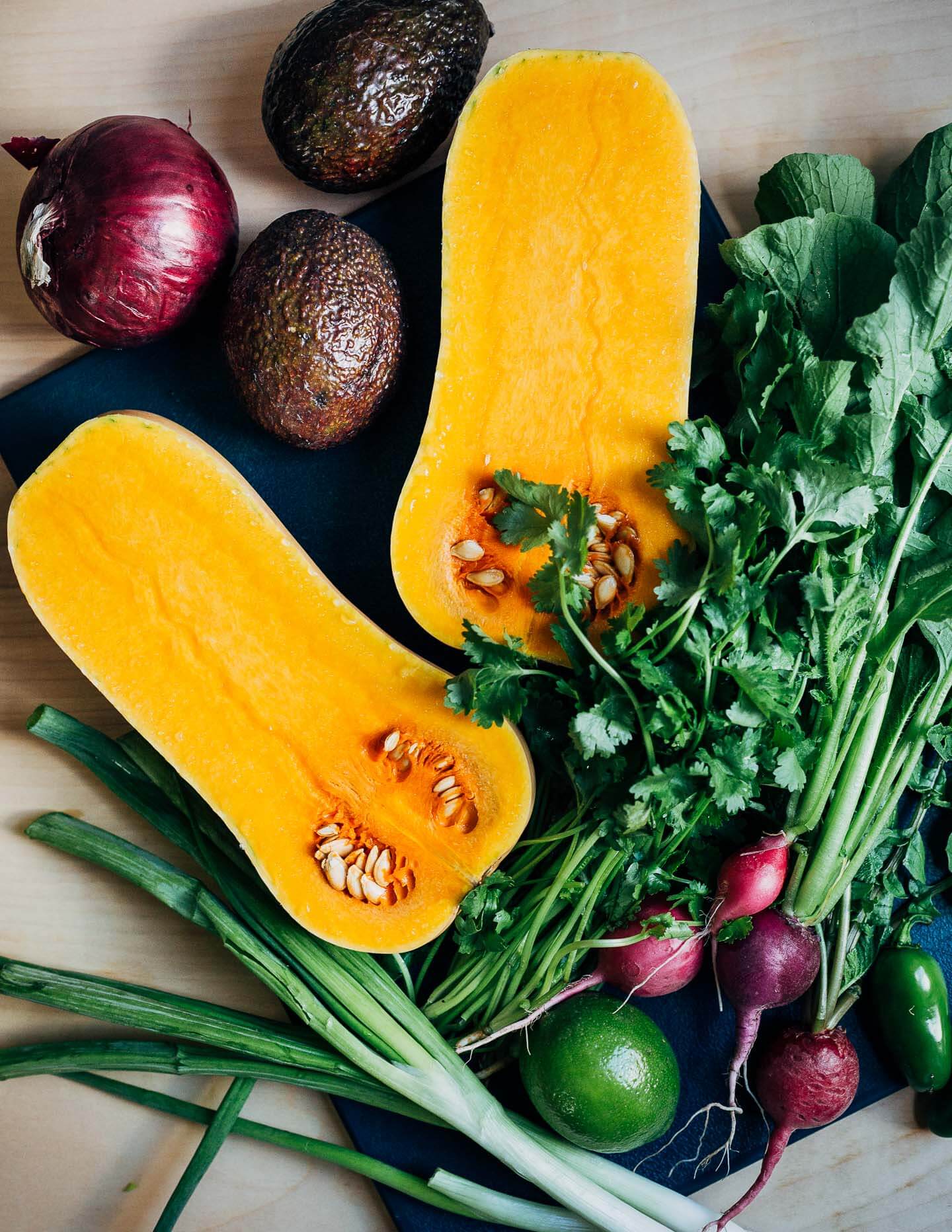 A halved butternut squash on a cutting board, along with greens, avocados, limes, and radishes. 