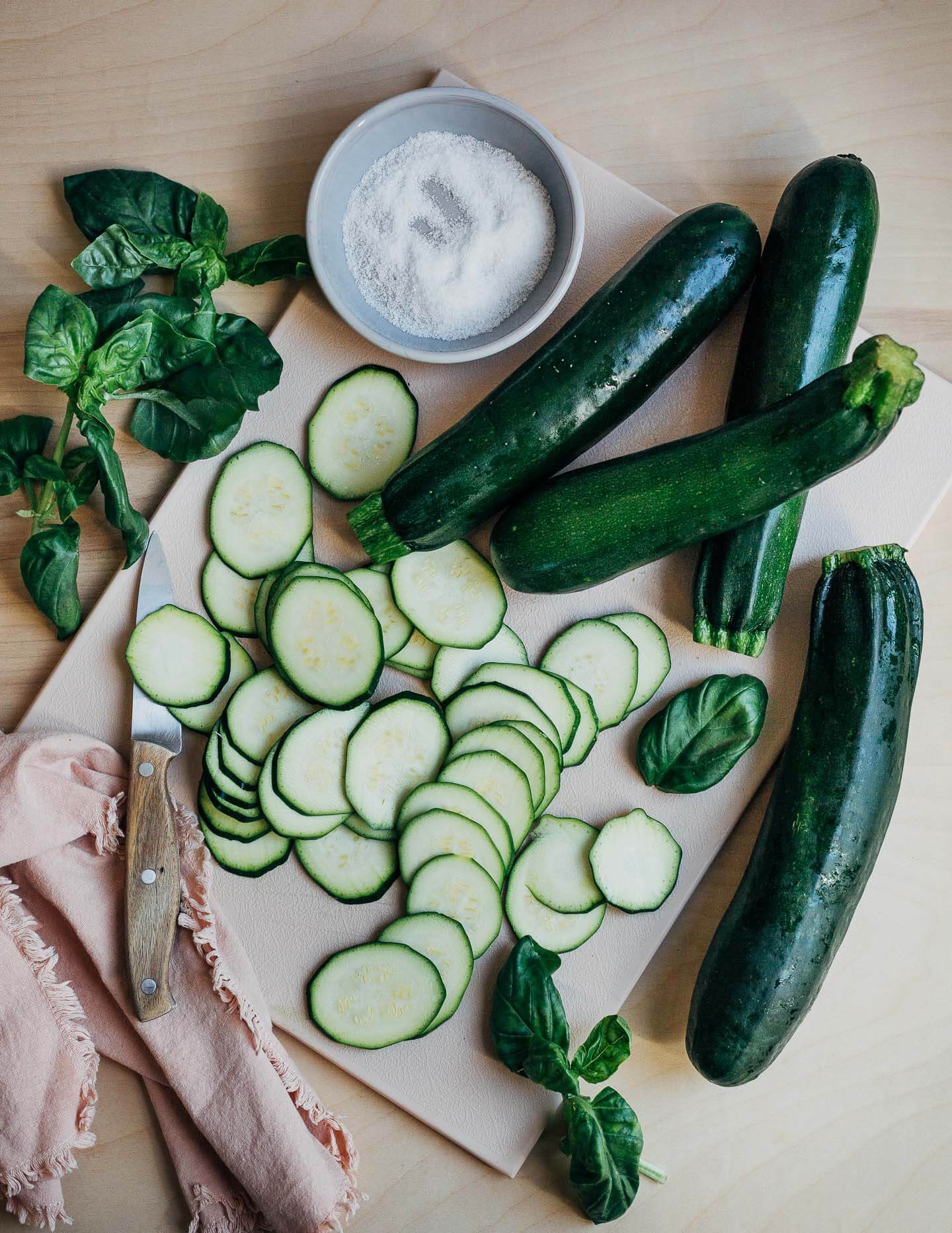 Whole and sliced zucchini on a cutting board with salt and basil on the side. 