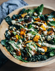 Kale salad in a wooden bowl with roasted squash cubes and a creamy dressing drizzle.