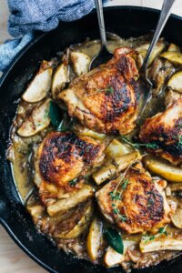 A cast iron skillet with golden chicken, apples, and herbs.