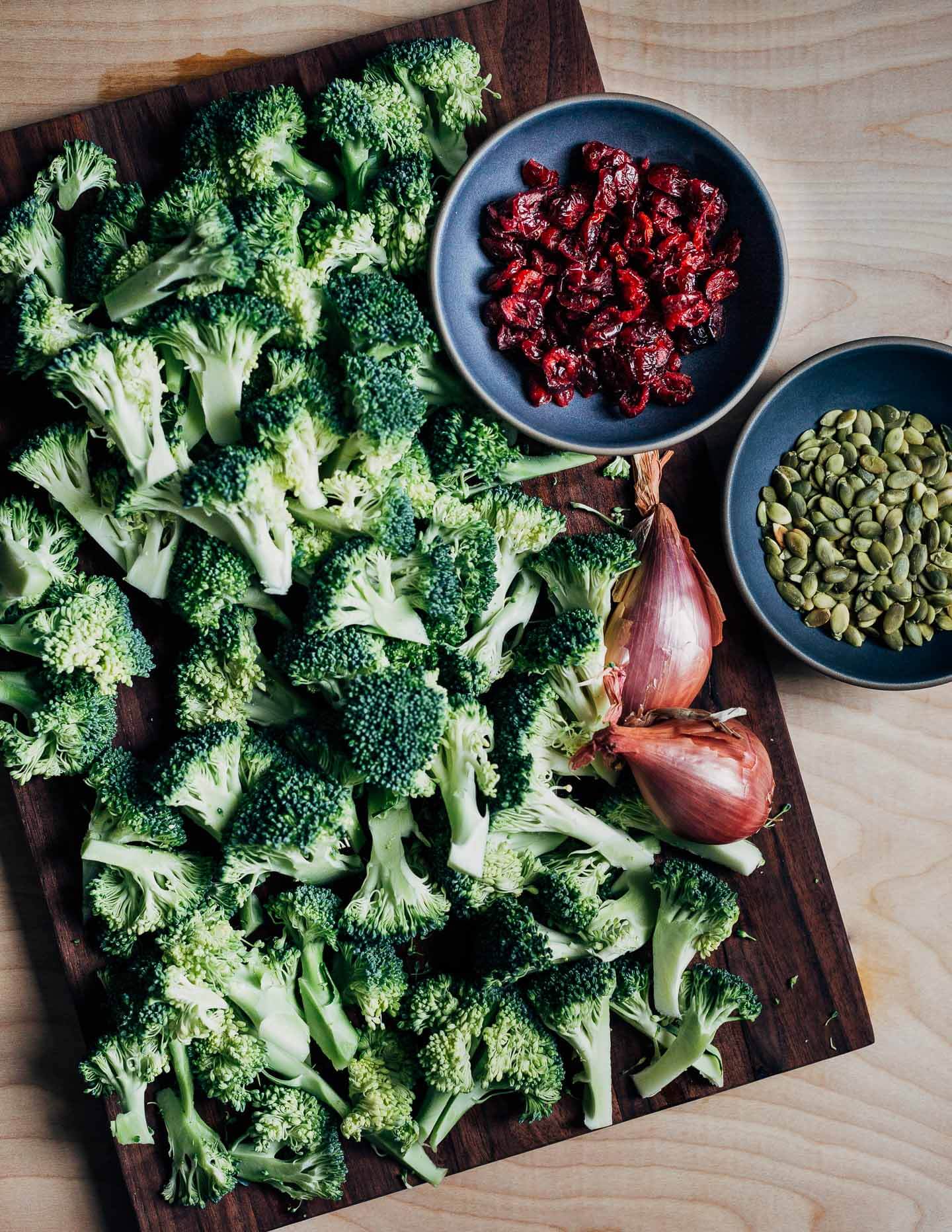 A cutting board with broccoli florets and shallots. Bowls of pumpkin seeds and cranberries are alongside. 