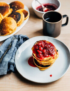 A stack on pancakes on a plate with a mug of tea and a pancake platter in the background.