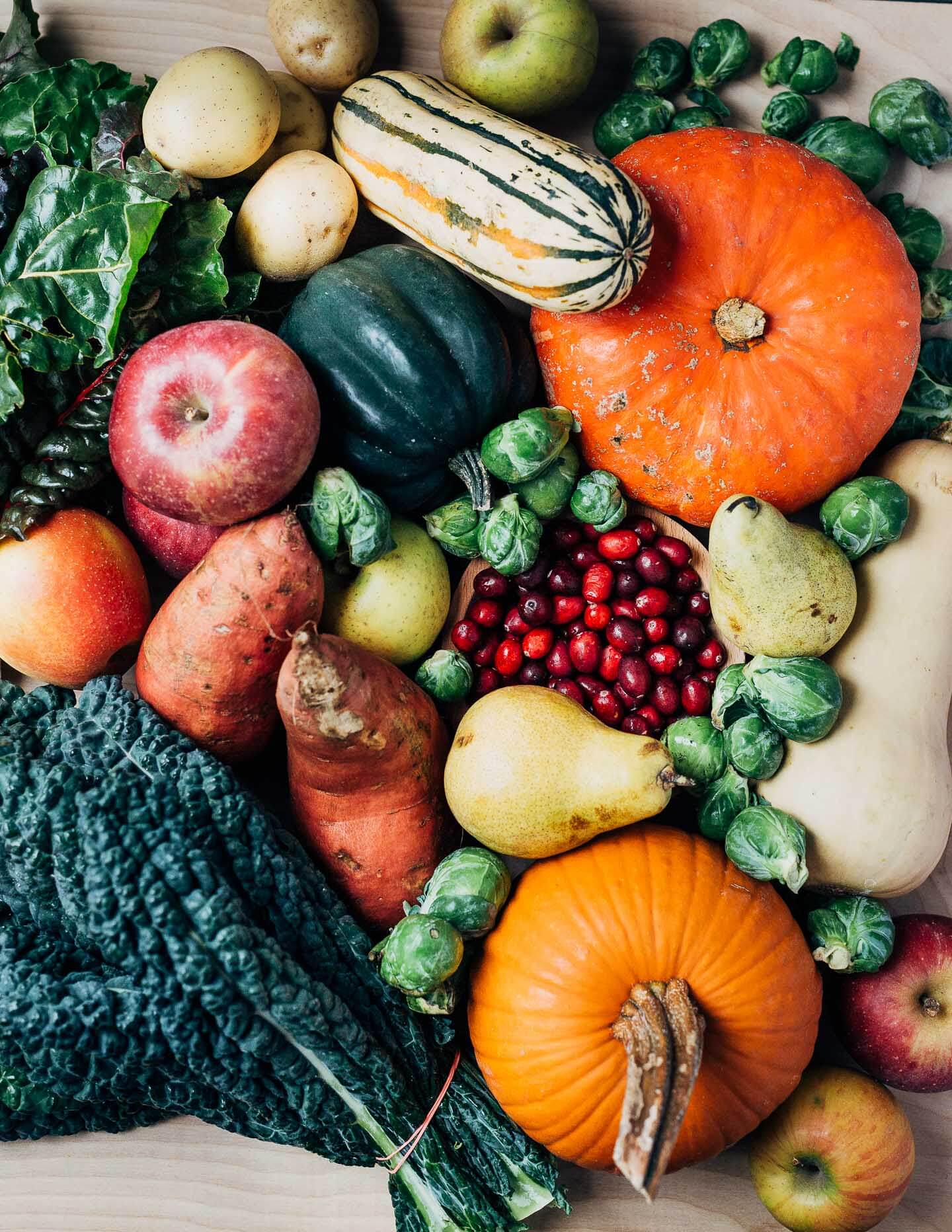 Produce piled on a table with squash, pumpkins, cranberries, Brussels sprouts, apples, pears, and greens. 