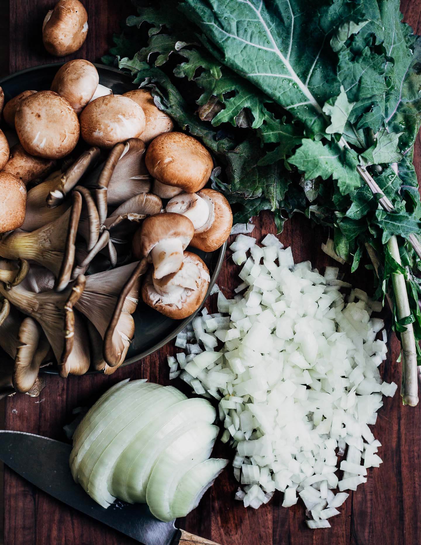 A cutting board piled with mushrooms, greens, and a partially chopped onion.