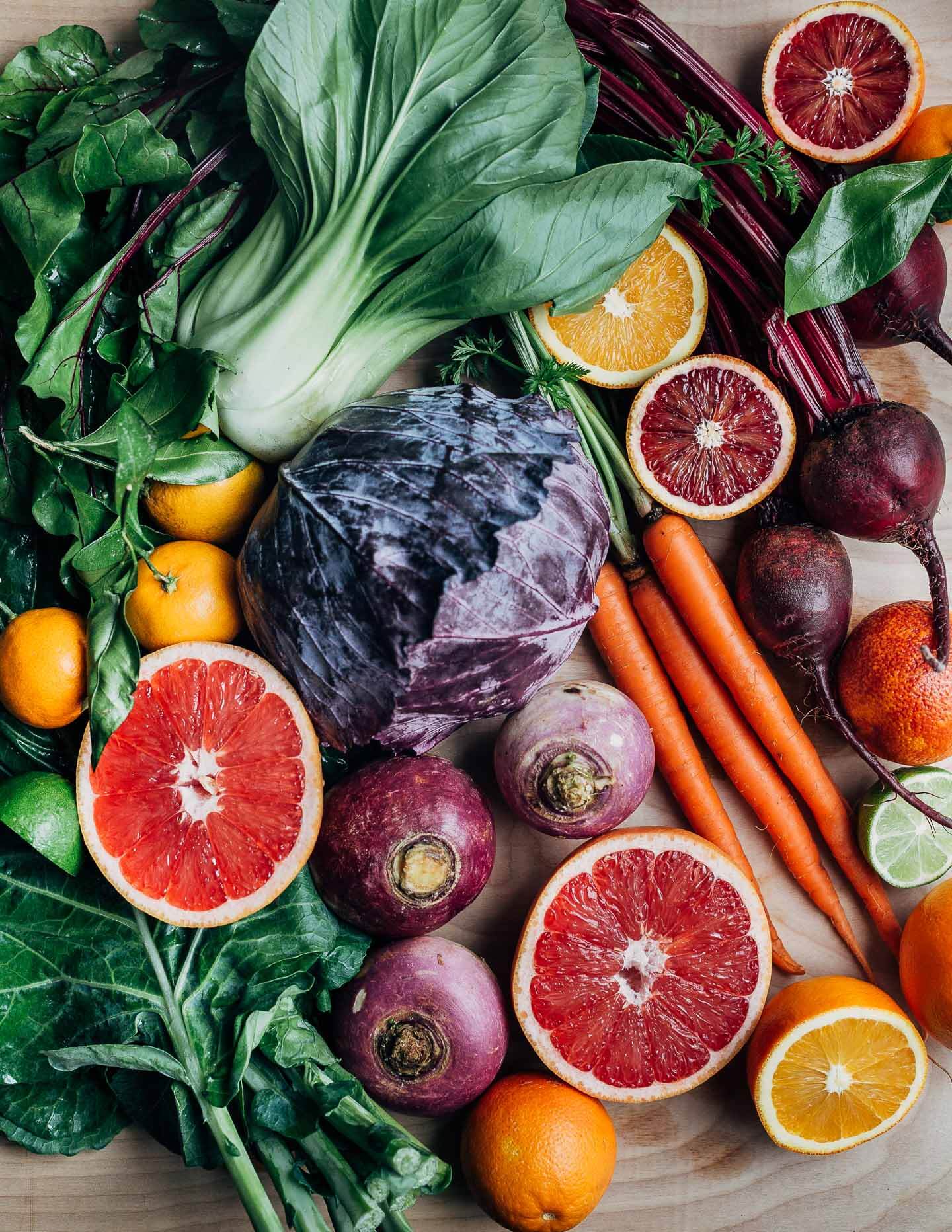 An assortment of fruits and vegetables arranged on a table, including bok choy, beets, carrots, grapefruit, and turnips. 