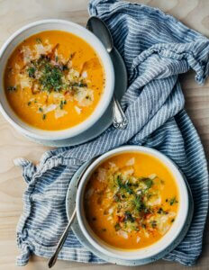 Two bowls of carrot soup with olive oil drizzled on top, along with herbs and Parmesan cheese.
