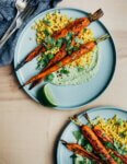 Two plates with roasted carrot, green tahini, and couscous. There are herbs and lime wedges scattered around the plates.
