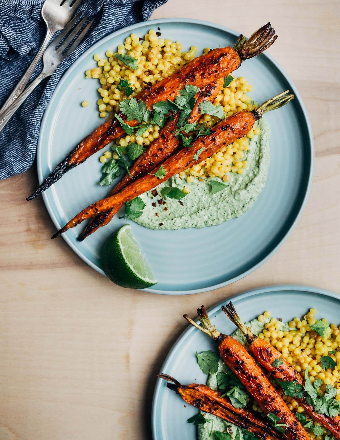 Two plates with roasted carrot, green tahini, and couscous. There are herbs and lime wedges scattered around the plates. 